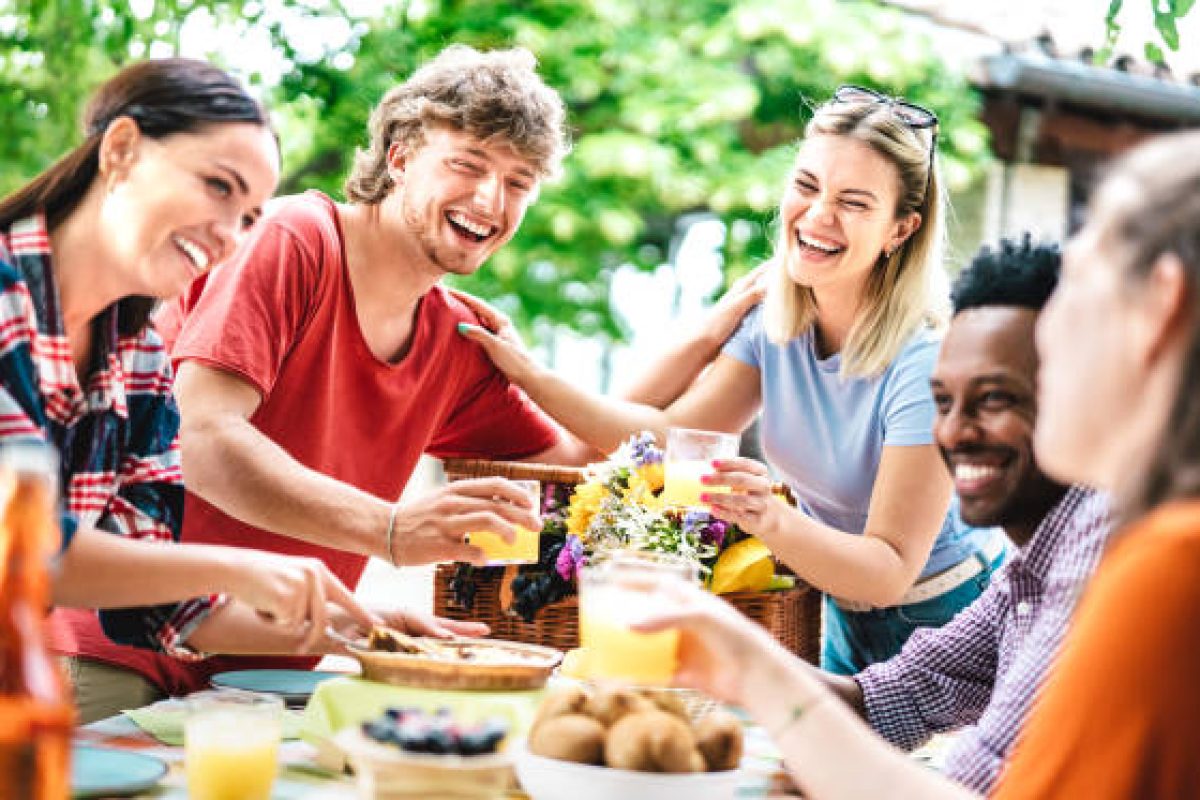 Happy young men and women toasting healthy orange fruit juice at farm house picnic - Life style concept with alternative friends having fun together on afternoon relax time - Bright vivid filter